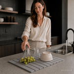 Woman in a linen shirt setting a glass to dry on the stone drying mat by the sink, with grapes and a ceramic bowl on the slats