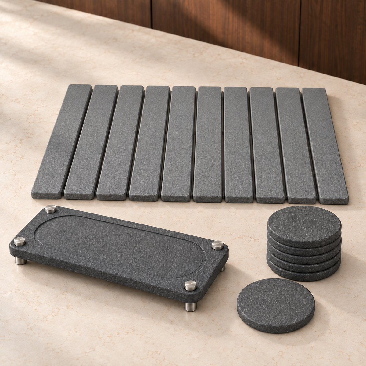 The three Essential Kitchen Set pieces on a stone counter: slatted stone drying mat, stone sink caddy on stainless steel legs and six stacked stone coasters